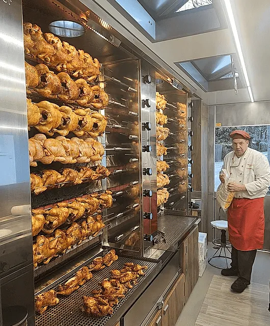 Mehrere Reihen von ganzen Hähnchen, die auf Drehspießen in einem großen Edelstahlgrill garen, mit einem Koch in weißer Jacke und roter Schürze daneben
