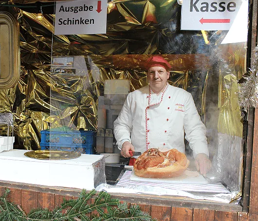 Mann in weißer Kochjacke hinter Theke mit großem Schinken, Schilder 'Ausgabe Schinken' und 'Kasse' hängen darüber